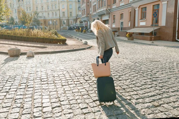 Молодая улыбающаяся блондинка с дорожной сумкой и телефоном, идущая по городской улице, женщина с длинными кудрявыми волосами, вид сзади — стоковое фото