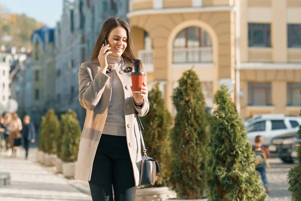 Young smiling beautiful woman walking on the street of a sunny autumn city talking on mobile phone — Stock Photo, Image