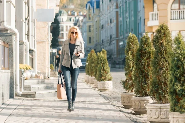 Молодая улыбающаяся блондинка с вьющимися волосами со смартфоном в руках, идущая по городской улице, осенний солнечный день — стоковое фото