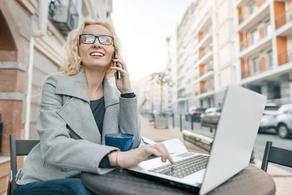Νεαρή όμορφη ξανθιά επιχειρηματίας με γυαλιά σε ζεστά ρούχα που κάθονται σε ένα υπαίθριο καφέ με φορητό υπολογιστή πίνοντας φλιτζάνι καφέ και μιλώντας στο τηλέφωνο, ιστορικό δρόμο της πόλης — Φωτογραφία Αρχείου