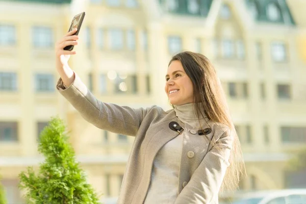 Outdoor portret uśmiechniętej szczęśliwej młodej kobiety z smartphone, miasto ulicy tle, jesienią słoneczny dzień. Dziewczyna robi zdjęcie selfie na smartfonie — Zdjęcie stockowe