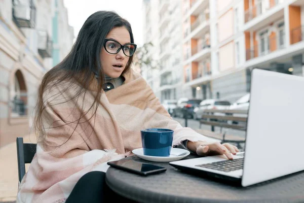 Νεαρή γυναίκα blogger ελεύθερη επαγγελματίας με ζεστά ρούχα σε υπαίθριο καφέ με φορητό υπολογιστή, κινητό τηλέφωνο. Αστικό υπόβαθρο — Φωτογραφία Αρχείου