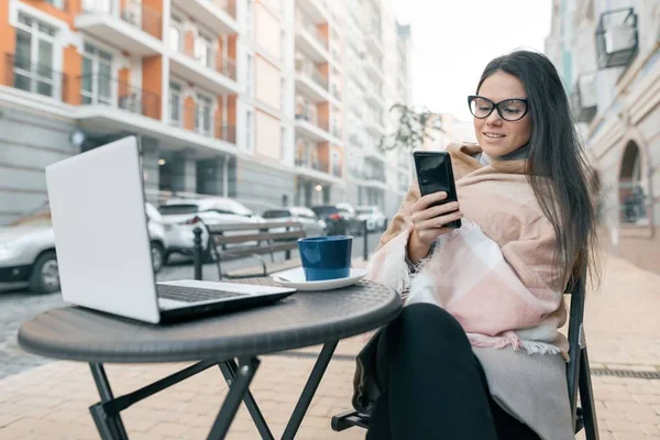 Jeune blogueuse pigiste en vêtements chauds dans un café extérieur avec ordinateur portable, téléphone mobile. Contexte urbain — Photo