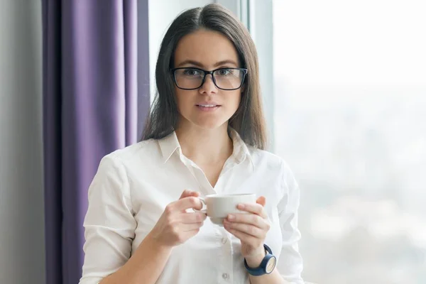 Porträtt av unga affärskvinna i vit skjorta bär glasögon med kopp kaffe, poserar leende kvinna tittar in i kameran, kontor i skyskrapa, molnigt dag — Stockfoto