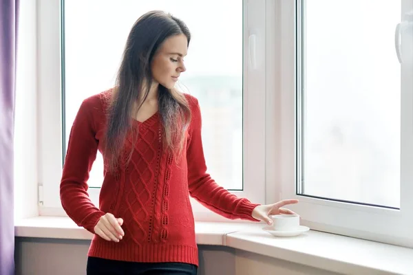커피를 마시는 빨간 스웨터에 젊은 아름다운 여자의 초상화입니다. 흐린 날 아침에 마천루의 창문 근처에 서 있는 여성 — 스톡 사진