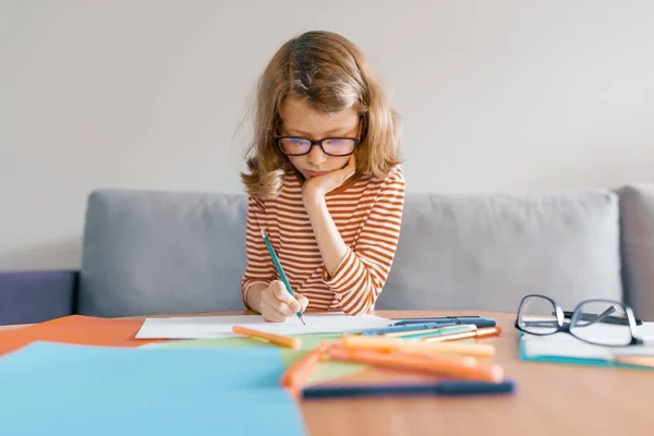 La muchacha de 8 años se sienta en el sofá en casa dibuja escribe con el lápiz sobre el papel. Rubia infantil con gafas estudiando en casa — Foto de Stock