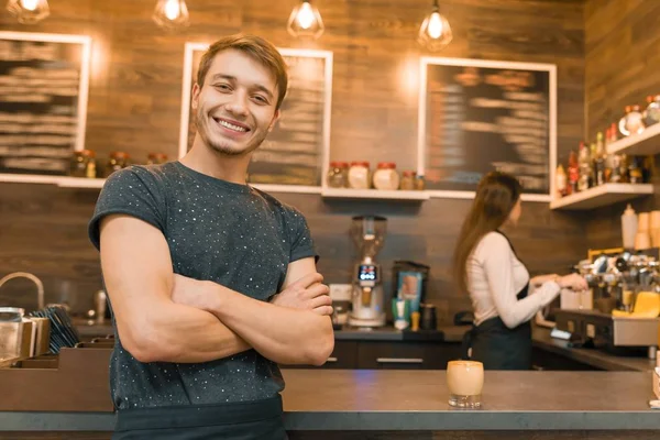 年轻的咖啡店男工微笑着看着摄像机，双臂交叉在吧台附近 — 图库照片
