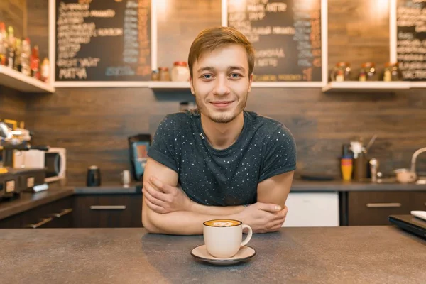 Portret van jonge lachende mannelijke café werknemer, staande bij de balie. Man met gevouwen handen met vers bereide koffie — Stockfoto