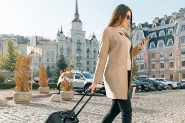 Genç güzel kadın seyahat bavul ve cep telefonu ile şehir sokak boyunca yürür. Moda esmer kız gülümseyen