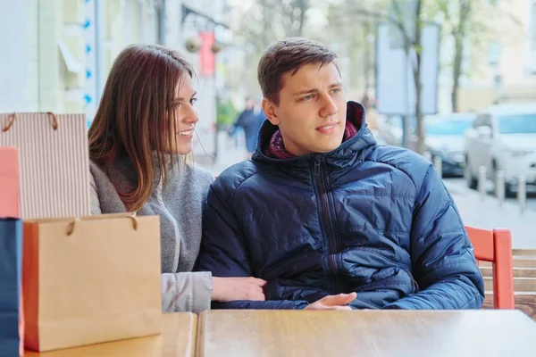 Young talking couple with shopping bags in street cafe, waiting for cup of coffee and tea