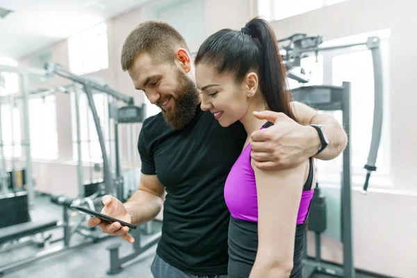 Mujer joven hablando con entrenador personal en el gimnasio, mirando en el teléfono inteligente y discutiendo. Fitness, deporte, entrenamiento, personas, concepto de estilo de vida saludable — Foto de Stock