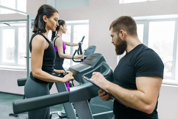 Jonge lachende fitness vrouwen met personal trainer een volwassen atletische man op de loopband in de sportschool. Sport, teamwork, training, gezonde levensstijl concept — Stockfoto