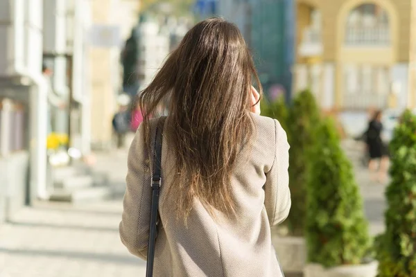 Młoda kobieta z telefonem komórkowym spacerująca po słonecznej ulicy miasta, dziewczyna ubrana w ciepły płaszcz, widok z tyłu, złota godzina — Zdjęcie stockowe