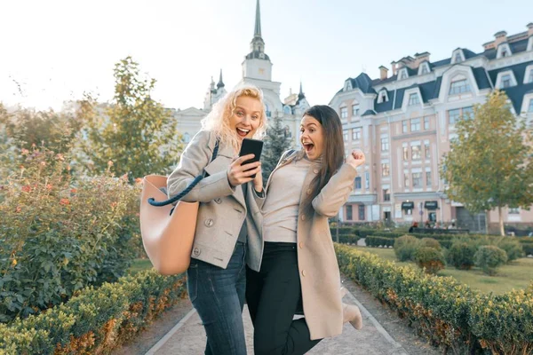 Dwie młode kobiety bawią się, patrząc na smartfon śmiech, słoneczny jesienny dzień, tło miasta, złotą godzinę — Zdjęcie stockowe