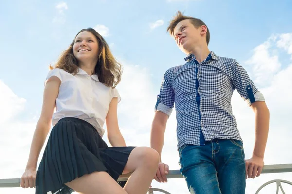 Happy couple of teens boy and girl 14, 15 years old. Young people smiling and talking, blue sky background