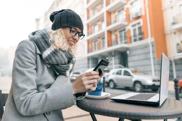 Jovem blogueira freelancer em chapéu de roupas quentes no café ao ar livre com computador portátil, telefone celular, cidade rua fundo — Fotografia de Stock