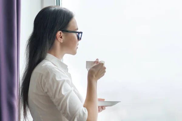 Retrato de mujer de negocios joven en gafas de camisa blanca con taza de café, mujer mirando por la ventana, oficina en rascacielos, día nublado — Foto de Stock