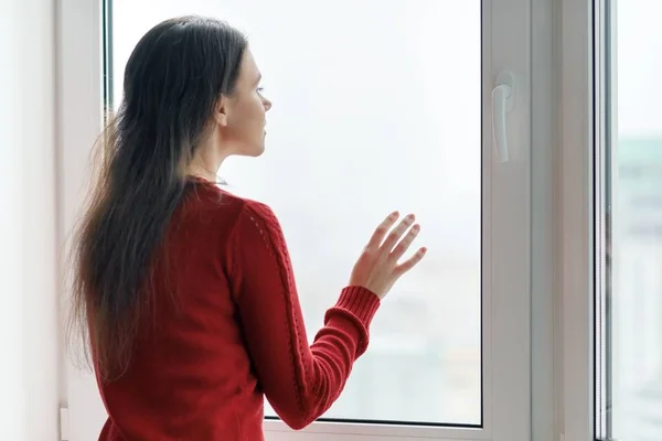 Mujer joven en suéter rojo mirando por la ventana, mujer puso sus manos en el vidrio de la ventana, vista lateral, ventana en rascacielos por la mañana en el día nublado — Foto de Stock