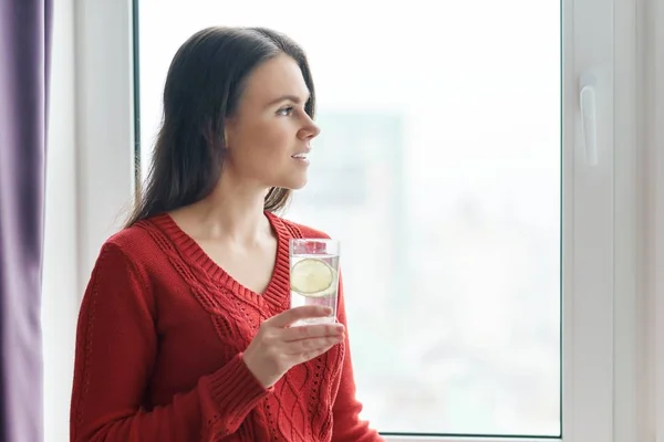 Joven hermosa mujer en suéter rojo con vaso de agua con cal, mujer está cerca de la ventana en rascacielos en el día nublado. Bebida saludable, antioxidante natural, vitaminas en la vida urbana — Foto de Stock