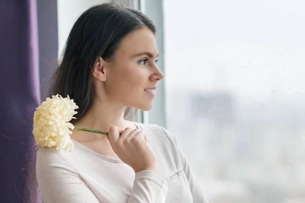 Portrait of young beautiful smiling woman with natural makeup, with large pale yellow flower in her hands, girl looking out the window with raindrops — Stock Photo, Image