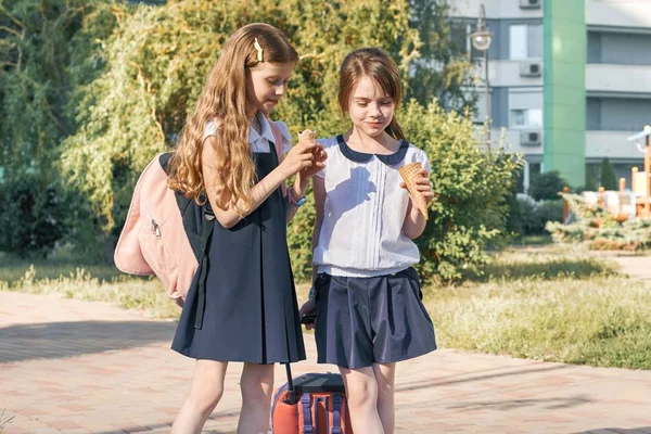 Ritratto all'aperto di due bambine con gli zaini in uniforme scolastica, sorridenti e che mangiano gelato — Foto Stock