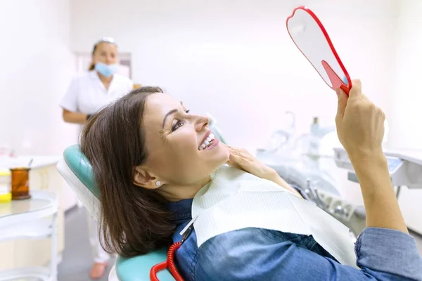 Proceso de tratamiento dental. Paciente femenina mirando sus dientes en el espejo mientras está sentada en la silla dental . — Foto de Stock