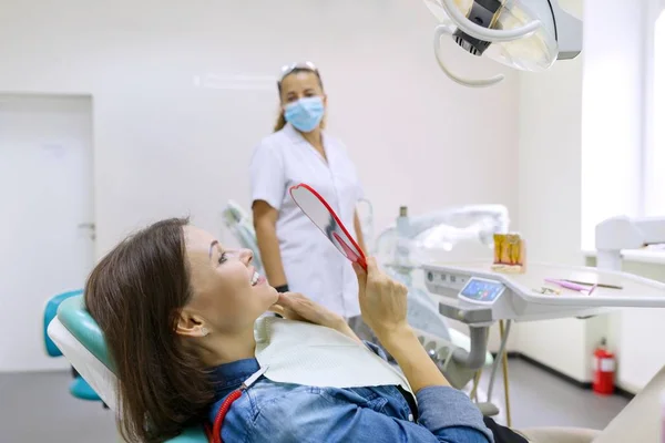 Process of dental treatment. Female patient looking at her teeth in mirror while sitting in dental chair. — Stock Photo, Image