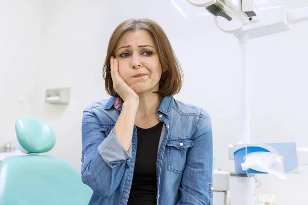 Mature woman holding hands over head with toothache, background dentist office.