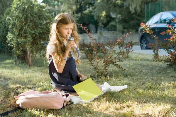女孩穿着校服，背包坐在院子里的草坪上，喝着瓶子里的水 — 图库照片