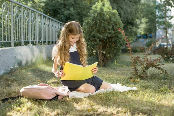 Retrato al aire libre de la colegiala sentada en el césped leyendo cuaderno de la escuela. Iniciar la escuela — Foto de Stock