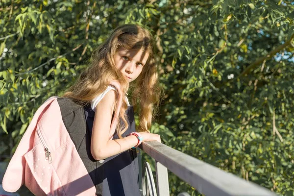 Chica colegiala rubia con mochila en uniforme escolar cerca de valla en el patio de la escuela —  Fotos de Stock