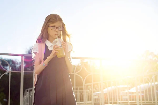 Outdoor portrait of girl elementary school student wearing glasses, school uniform, with backpack drinking natural juice from glass. — Stock Photo, Image