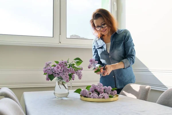Зрелая женщина делает букет из лиловых ветвей дома за столом — стоковое фото