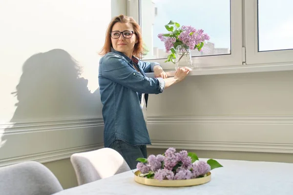 Portret van mooie rijpe vrouw thuis met boeket van lila bloemen. Achtergrond van Home interieur dineren in de buurt van het raam — Stockfoto