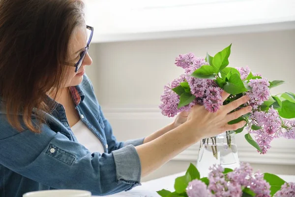 Middle-aged woman making bouquet of lilac branches, close-up female arranging flowers bunch in vase