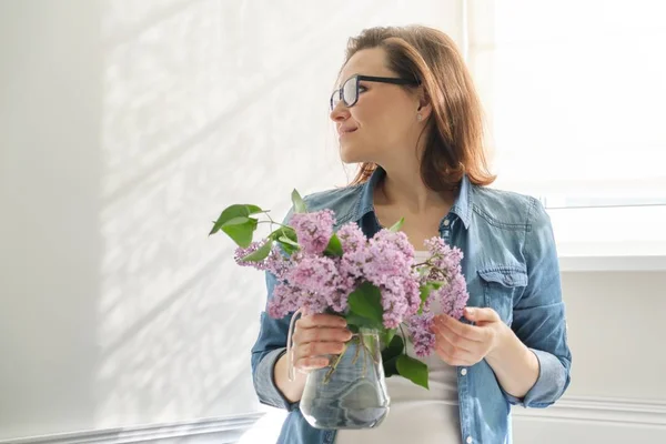 Porträt der schönen reifen Frau zu Hause mit einem Strauß fliederfarbener Blumen. Hintergrund des häuslichen Esszimmers am Fenster — Stockfoto