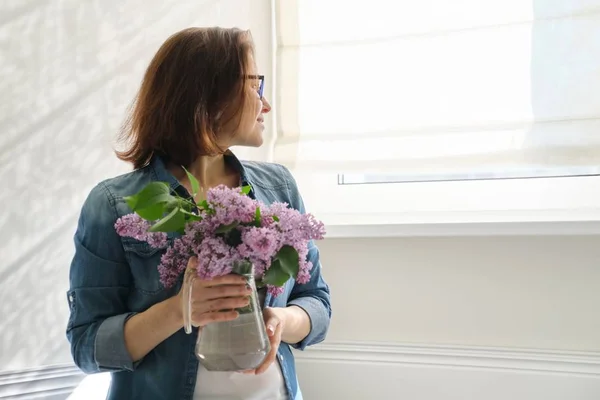 Retrato de una hermosa mujer de mediana edad en casa con ramo de flores lila. Luz de fondo pared luz solar en la ventana —  Fotos de Stock