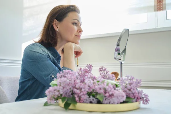 Rijpe vrouw 40 jaar oud kijkt naar haar gezicht in de spiegel, vrouw zittend aan de tafel thuis make Makeup — Stockfoto