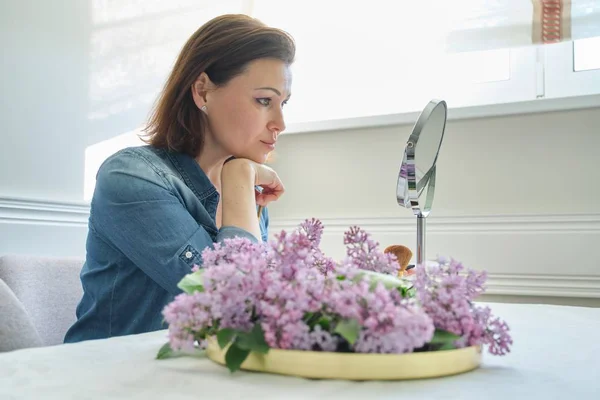 Rijpe vrouw 40 jaar oud kijkt naar haar gezicht in de spiegel, vrouw zittend aan de tafel thuis make Makeup — Stockfoto
