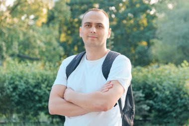 Olgun gülümseyen pozitif adam Açık portre. Kameraya bakan katlanmış elleri ile sırt çantası ile beyaz t-shirt kendinden emin erkek