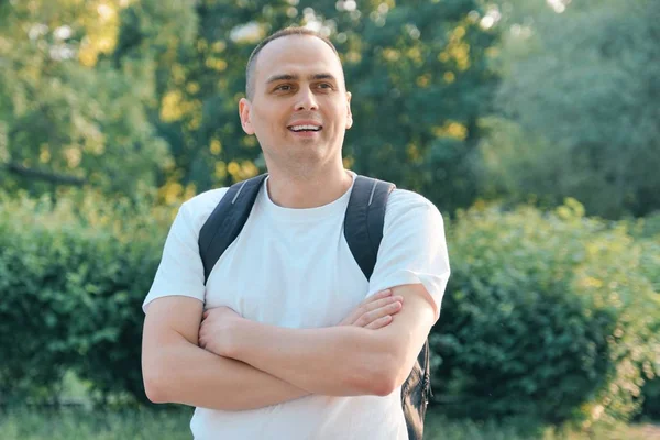 Olgun gülümseyen pozitif adam Açık portre. Katlanmış elleri ile sırt çantası ile beyaz t-shirt kendinden emin erkek