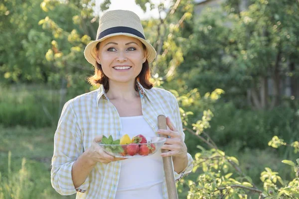 Retrato al aire libre de mujer madura positiva en sombrero de paja. Sonriente hembra con plato de fresas menta limón, fondo verde jardín, día soleado puesta de sol — Foto de Stock