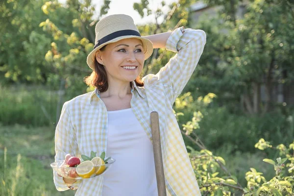Utomhus porträtt av lycklig kvinna 40 år gammal, hona i trädgården i strå hatt med tallrik jordgubbar Mint citron — Stockfoto