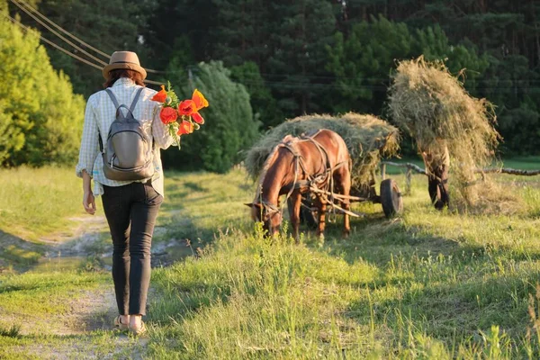 Donna in cappello con mazzo di fiori di papaveri rossi passeggiando lungo la strada di campagna rurale, vista posteriore. Fondo rustico, cavallo con erba — Foto Stock