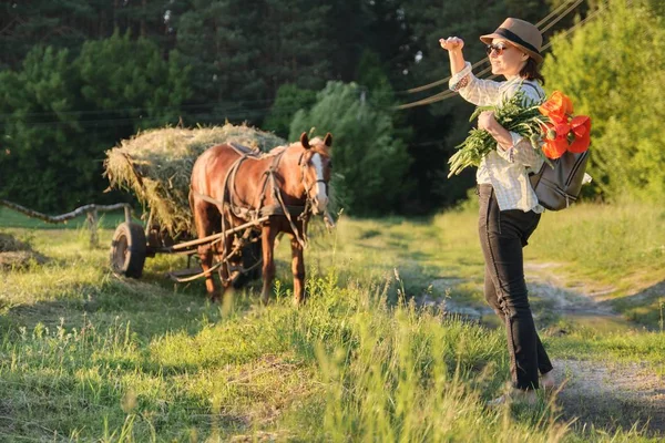 Stile rustico, donna matura felice in cappello con mazzi di fiori papaveri passeggiando lungo la strada di campagna — Foto Stock