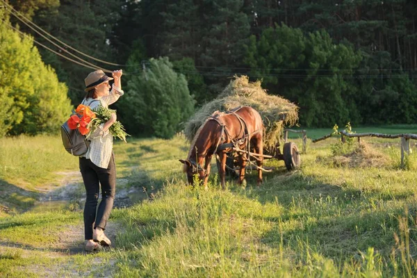 Frau mit Hut mit Strauß roter Mohnblumen, die ländliche Landstraße entlang geht, Rückansicht. rustikaler Hintergrund, Pferd mit Gras — Stockfoto