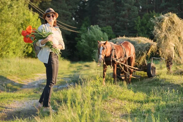 Stile rustico, donna matura felice in cappello con mazzi di fiori papaveri passeggiando lungo la strada di campagna — Foto Stock