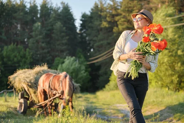 Rustik stil, Mogen lycklig kvinna i hatt med buketter av vallmo blommor promenader längs landsväg — Stockfoto