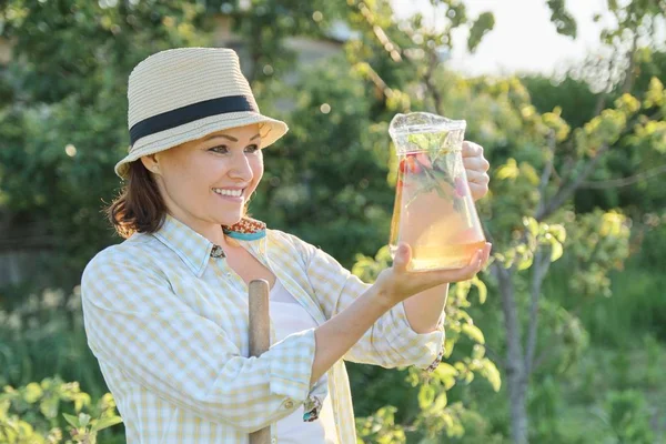 Porträtt av mogen lycklig kvinna i hatt i trädgården med naturliga hemlagad ört dryck med mynta jordgubbar — Stockfoto