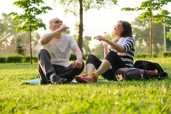 Ευτυχισμένο ώριμο ζευγάρι που κάθεται στο πάρκο στο χαλάκι γυμναστικής, πίνοντας γιαούρτι μετά από αθλητικές ασκήσεις. — Φωτογραφία Αρχείου